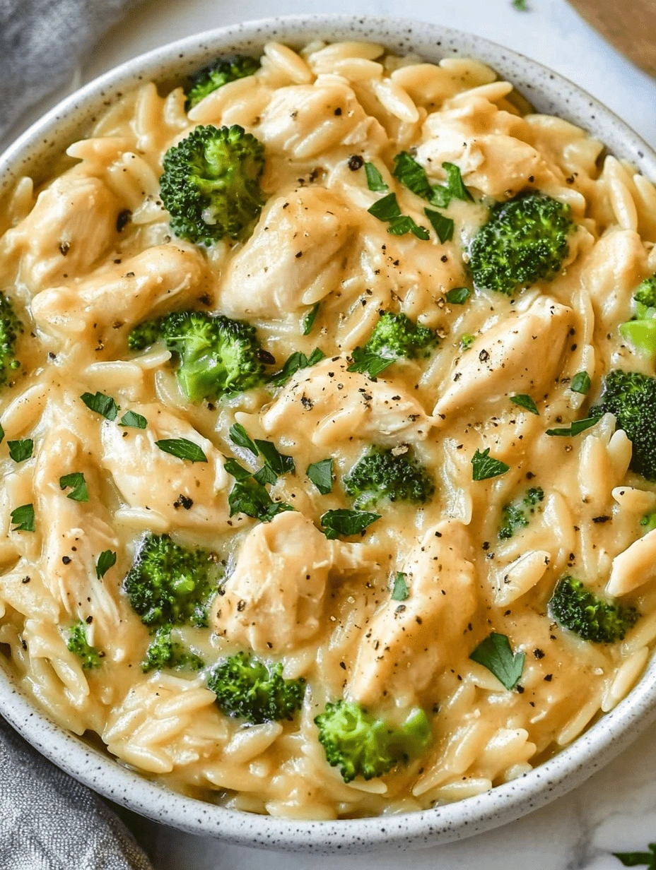 Top-down shot of a white bowl filled with cheesy chicken broccoli orzo pasta, garnished with parsley and black pepper.